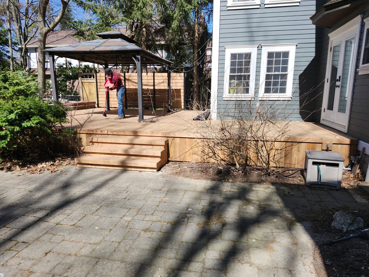 Residential backyard with wooden deck, paver patio, shade structure, and gray house with windows