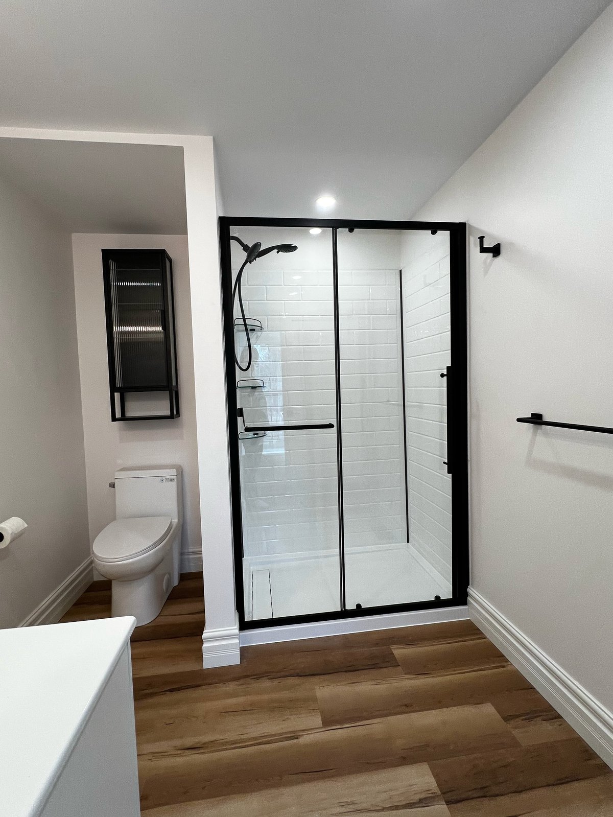 Modern bathroom with black-framed glass shower enclosure, white subway tile walls, wooden flooring, white vanity, and toilet