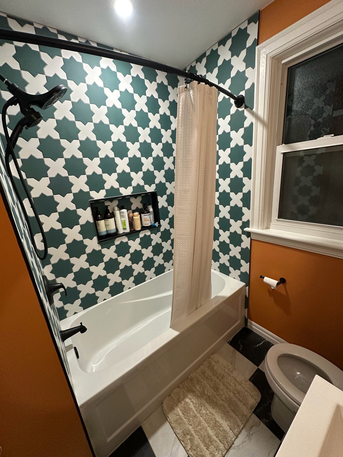 Modern bathroom with teal and white patterned tile walls, white bathtub with shower curtain, toilet, and burnt orange accent wall