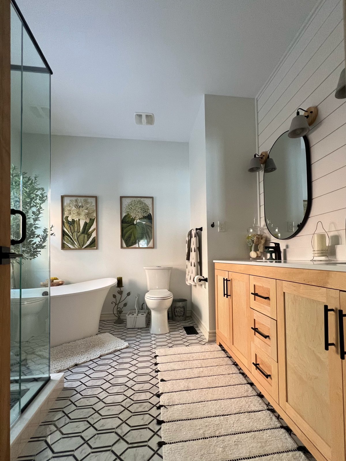 Modern bathroom with wooden vanity, hexagonal floor tiles, freestanding tub, glass shower enclosure, and botanical wall art