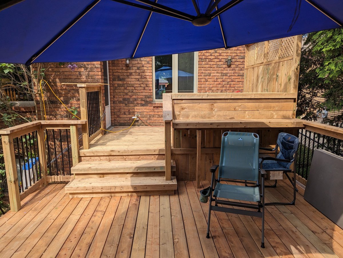Wooden deck with chairs and blue umbrella attached to a brick house with stairs and railing
