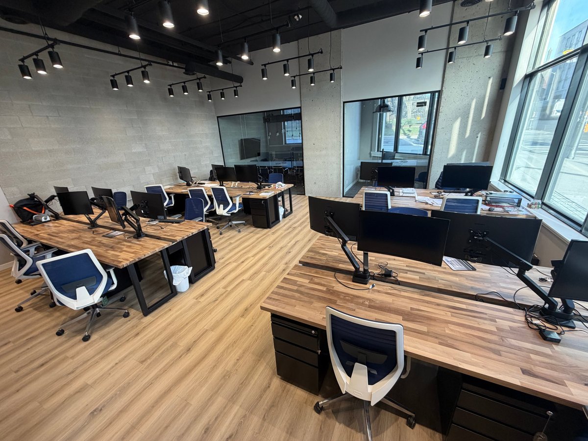 Modern office space with wooden desks, black ergonomic chairs, computer monitors, and floor-to-ceiling windows with natural lighting