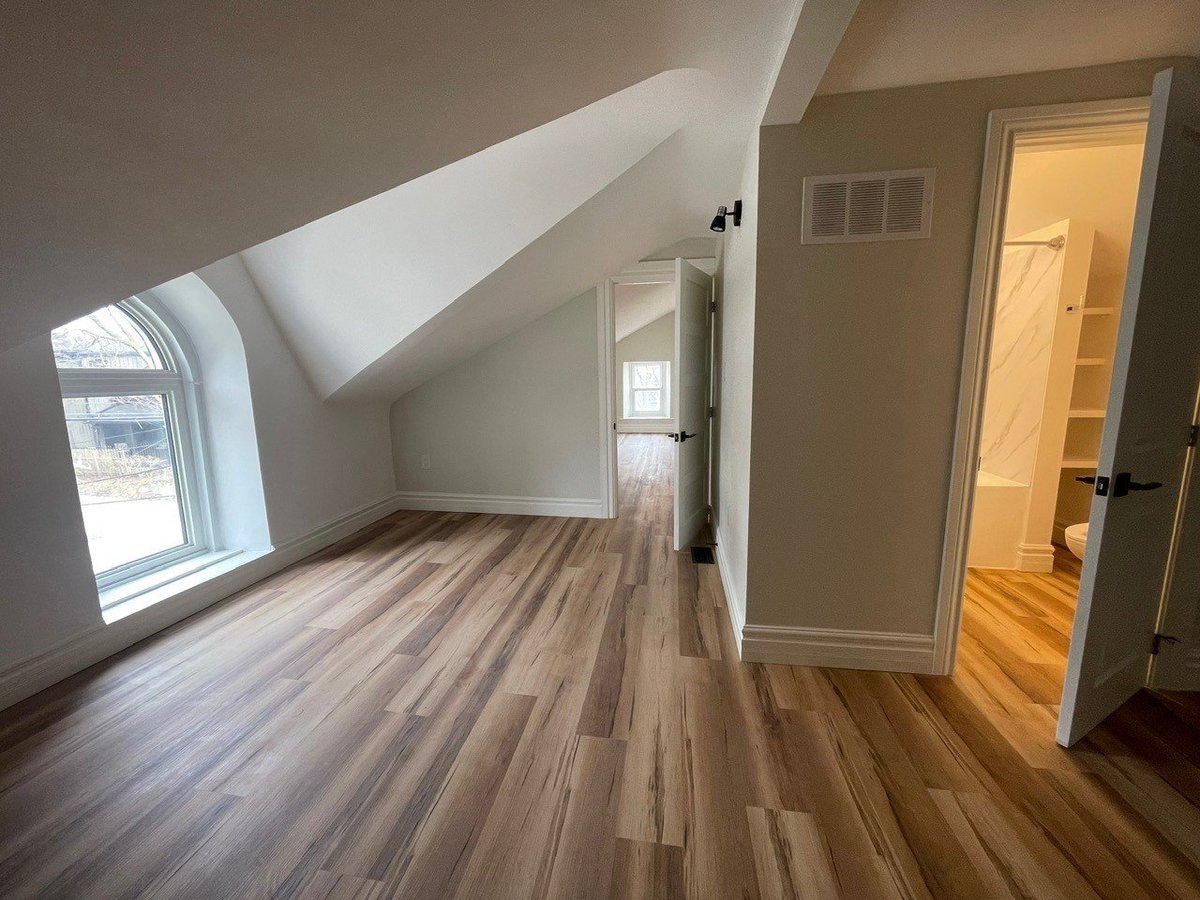 Empty attic room with wood flooring, sloped ceiling, arched window, and doorway to hallway