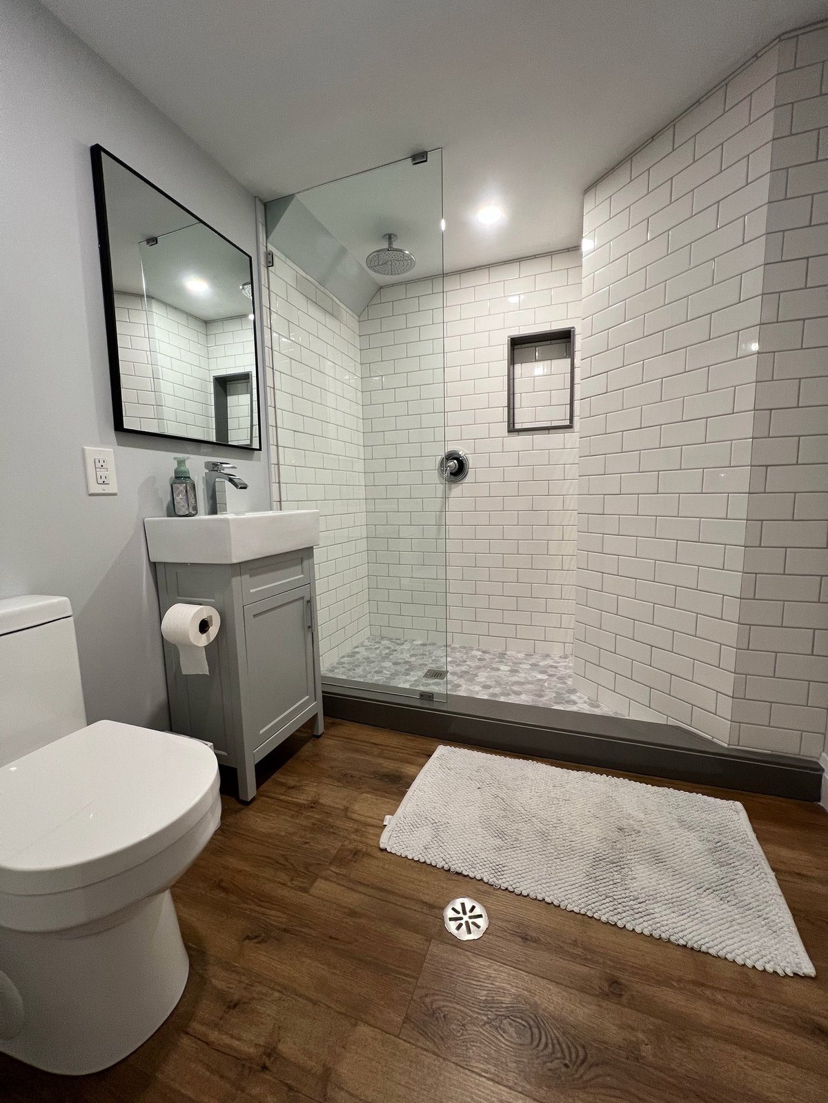 Modern bathroom with white subway tile shower, gray vanity, framed mirror, toilet, and wooden flooring
