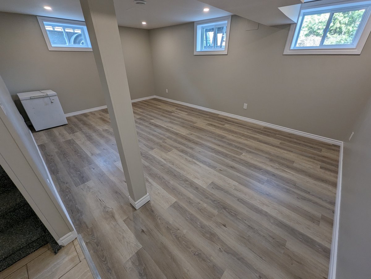Empty basement room with laminate wood flooring, white walls, support columns, windows, and recessed lighting