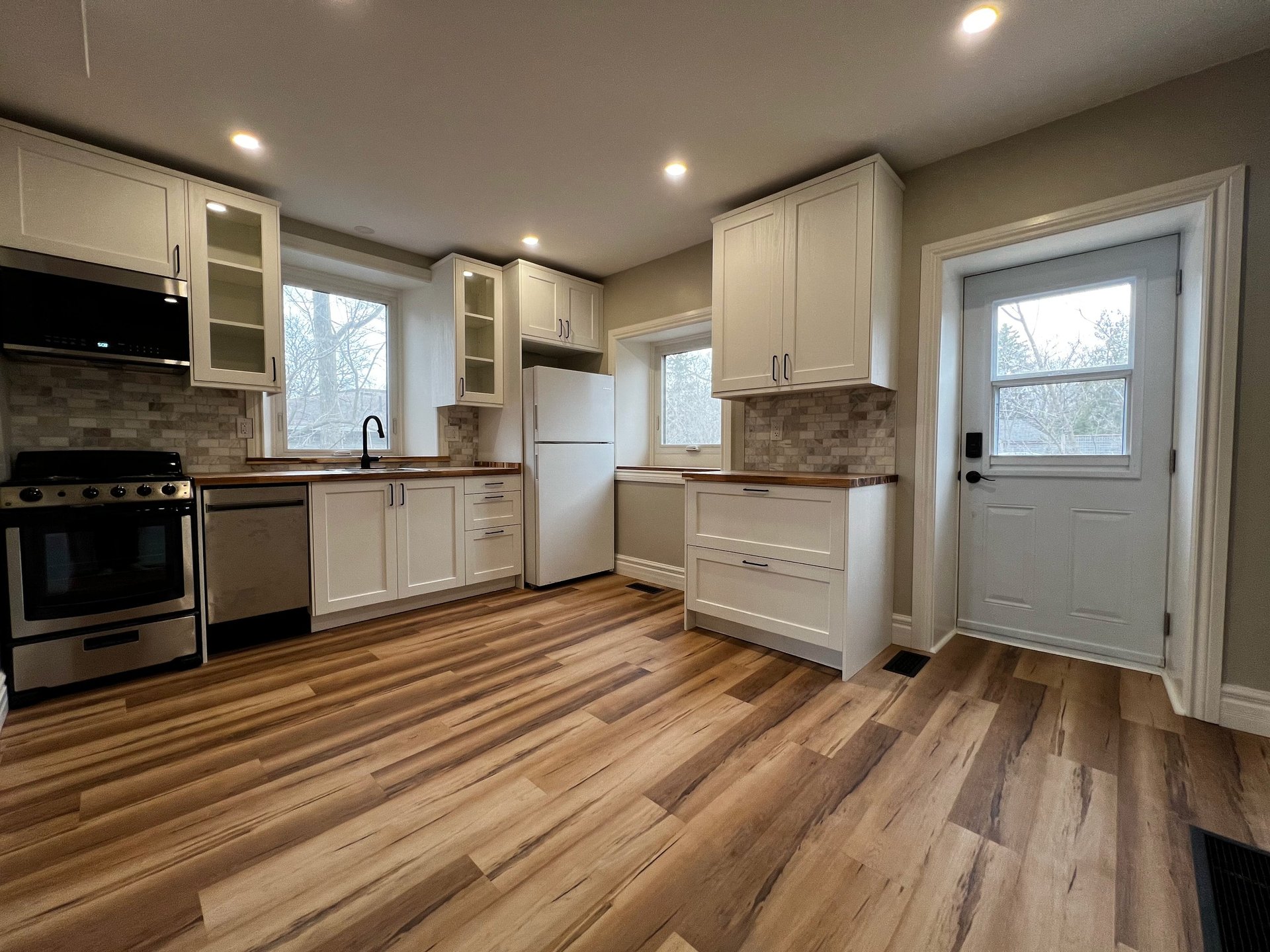 Modern kitchen with cream cabinets, stainless steel appliances, wood-grain flooring, and windows overlooking natural landscape