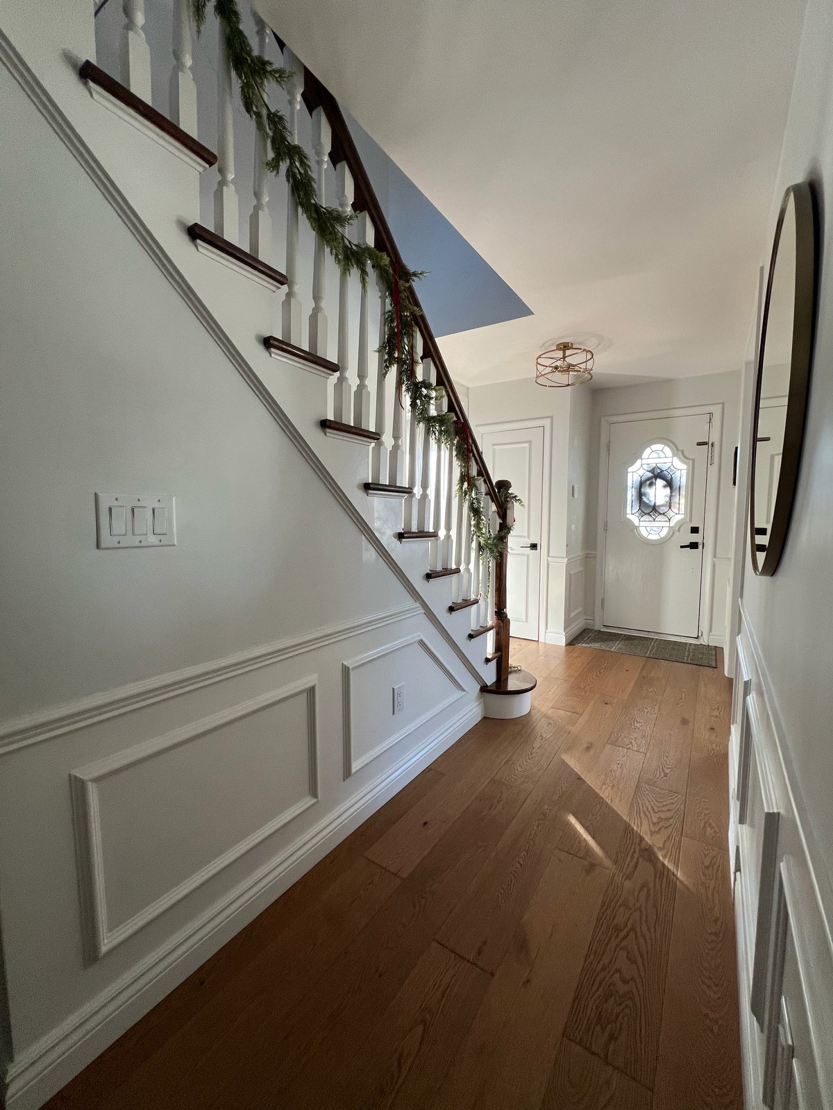 Elegant entryway with wooden staircase decorated with garland, wainscoting, and hardwood floors leading to front door