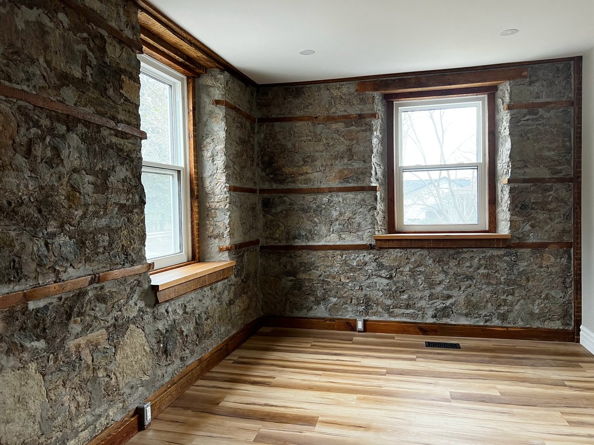 Empty room with exposed stone walls, wooden flooring, and two windows during renovation